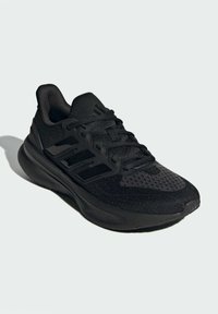 Chaussure de sport noire en maille et en matériaux synthétiques. Elle possède une surface texturée, une semelle rembourrée et un design à trois bandes sur le côté.