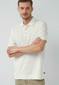 Biely polo tričko s krátkym rukávom a tromi zlatými gombíkmi, rebrovaná textúra a rovný lem. Malá značka na dolnej strane.