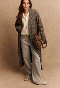 Mujer vestida con un abrigo largo de cuadros, un cárdigan crema, jeans de pierna ancha y planos beige, sosteniendo un bolso de ante marrón contra un fondo neutro.