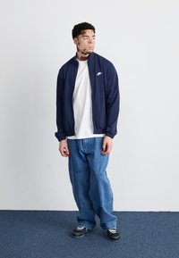 Casaco azul-marinho com fecho éclair e punhos elásticos sobre uma t-shirt branca, combinado com jeans azuis largos e sapatilhas pretas com detalhes brancos.