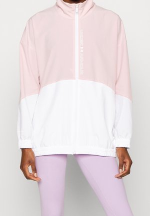 Osoba nosící dvoubarevnou růžovo-bílou bundu na zip s logem "Under Armour" na hrudi, doplněnou světle fialovými legínami.