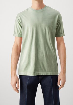 Homme portant un t-shirt vert clair à manches courtes avec manches retroussées et un jean bleu foncé, debout devant un fond uni.