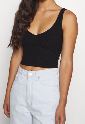 Kobieta ubrana w czarny prążkowany, bezrękawnikowy krótki top i jasnoniebieskie, wysokowycięte dżinsy na prostym tle.