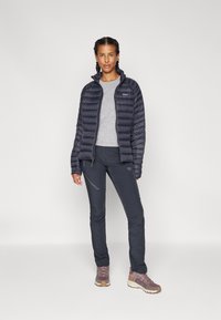 Marineblauwe pufferjack met verticale gestikte patronen, grijze longsleeve top, donkere broek en lichtbruine wandelschoenen, voor een effen achtergrond.