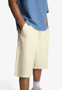 Pantaloni corti in lino beige chiaro con vita elasticizzata, tasche laterali e vestibilità rilassata, abbinati a un top blu con texture.