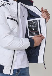 Chaqueta acolchada blanca con acentos en azul marino, cierre de cremallera y un bolsillo interior que muestra las instrucciones de cuidado y características en una etiqueta negra.
