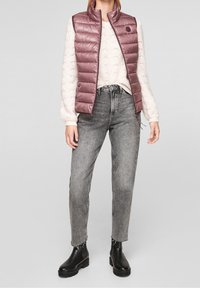 Chaleco acolchado lila claro con cuello alto, combinado con un suéter crema de punto trenzado y jeans grises de talle alto. Botines negros completan el conjunto.