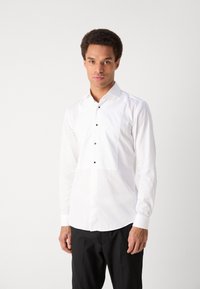 Fehér, hosszú ujjú ing, amely kiemelkedő négyzetes folttal rendelkezik a mellrészen, fekete gombokkal és klasszikus gallérral. Sima anyagtextúra.