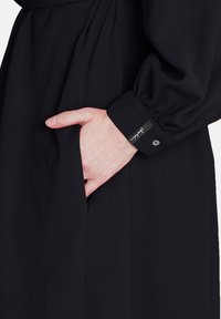 Rochie neagră cu mâneci bufante, având o mână în buzunarul lateral. Prezintă un manșon cu nasturi și un accent cu etichetă pe mânecă. Material neted.