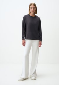 Felpa grigia scura con collo rotondo e maniche lunghe, abbinata a pantaloni bianchi a gamba larga con aperture laterali e sneakers bianche.
