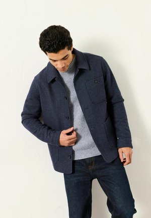 Marineblaue Jacke aus Wollmischung mit Kragen, vorderen Knöpfen und einer Brusttasche. Getragen über einem grauen Strickpullover und kombiniert mit dunklen Jeans.