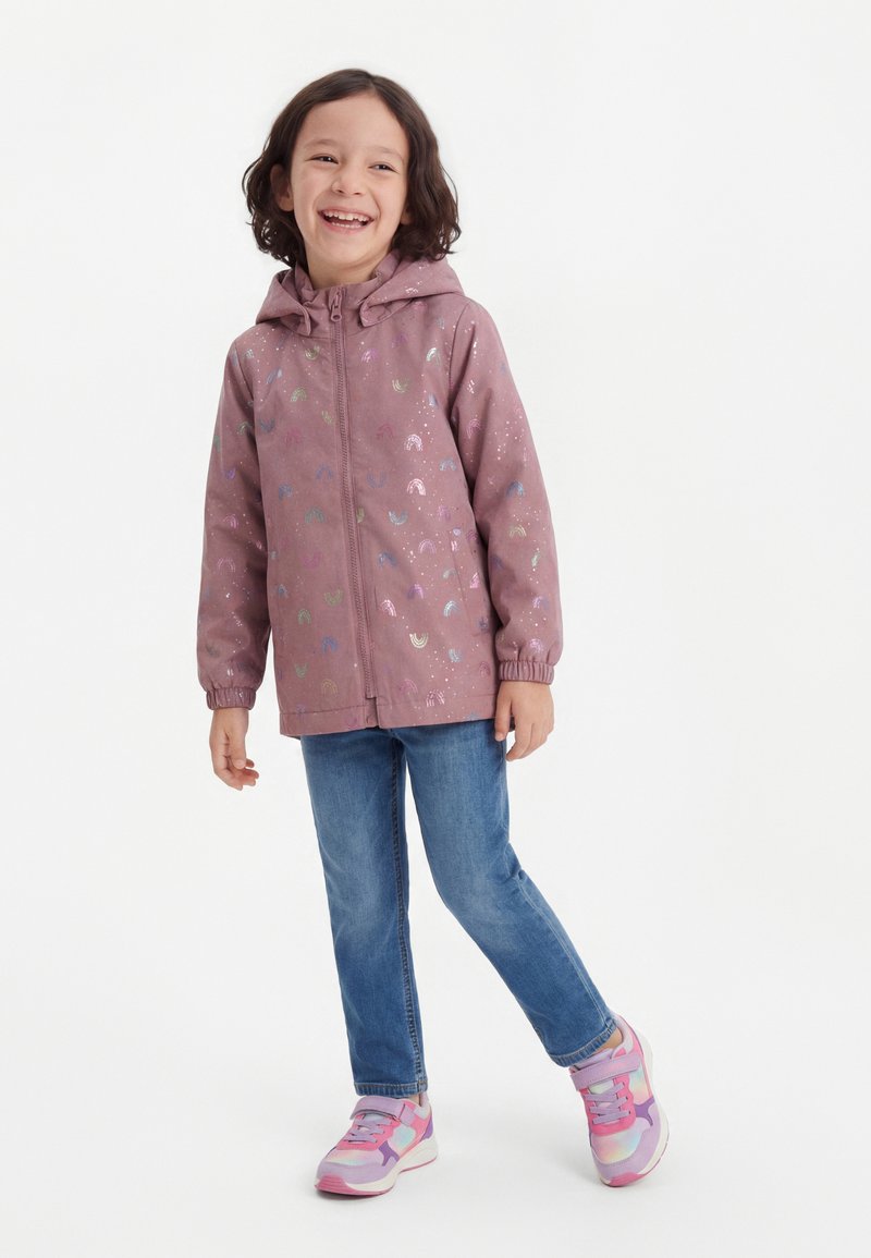 Enfant souriant portant une veste à capuche mauve avec des motifs arc-en-ciel, un jean bleu et des baskets roses, debout devant un fond blanc uni.