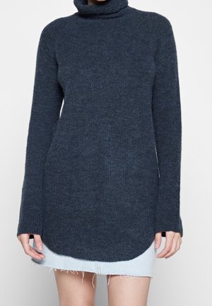 Femme portant un pull à col roulé gris foncé à manches longues avec un ourlet asymétrique sur une jupe en jean bleu clair effilochée.
