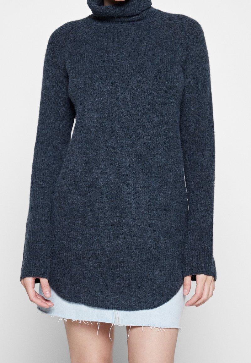 Femme portant un pull à col roulé gris foncé à manches longues avec un ourlet asymétrique sur une jupe en jean bleu clair effilochée.