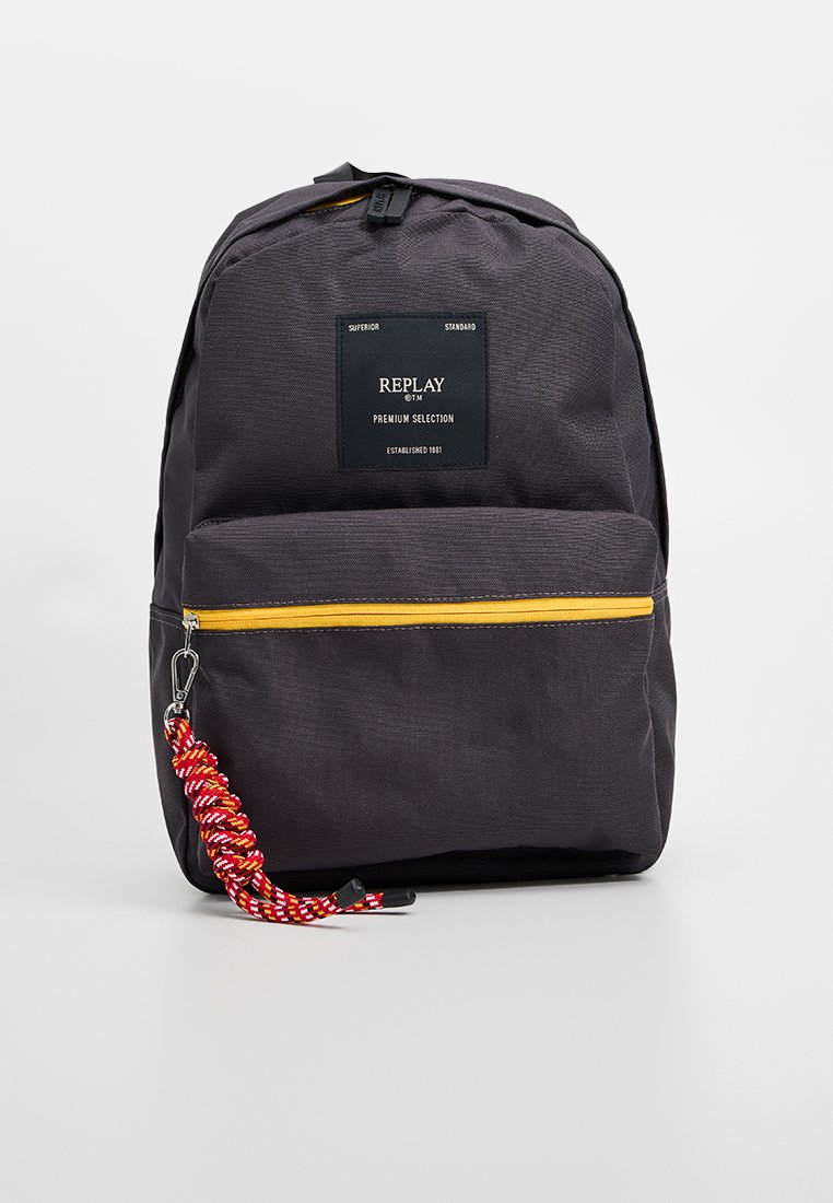 Zaino realizzato in tessuto nero testurizzato con una cerniera gialla, dotato di una tasca frontale e di un dettaglio con fascia intrecciata rossa e bianca.