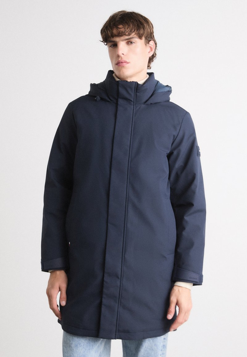 Marineblå, vandtæt parka med hætte, høj krave, frontlynlås og sidelommer. Har lange ærmer og kontrastfarvet indenfor.