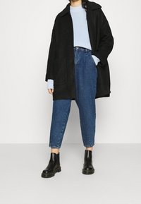 Musta ylipitkä takki, jossa on kaulus, vaaleansininen ribattu neule, siniset korkeavyötäröiset farkut ja mustat nilkkurit. Yksinkertainen ja rento muotoilu.