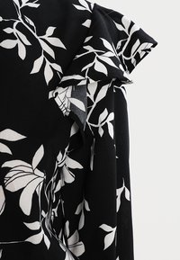 Tessuto nero con un motivo floreale bianco, con dettagli arricciati alla spalla. Texture liscia. Dettaglio ravvicinato del design della manica.