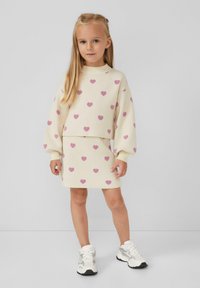 Ragazzina dai lunghi capelli biondi che indossa un completo di maglione e gonna color crema con cuori rosa e sneakers bianche, in piedi contro un muro grigio uniforme.