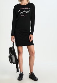 Svart långärmad tröja med "Forest Park Portland 1994" tryckt i vitt och rosa. Bärs med en svart bodycon-kjol och svarta sneakers; håller en svart ryggsäck.