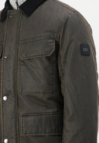 Giacca cerata marrone con colletto in corduroy nero, tasche a pattina sul frontale, bottoni a pressione in metallo e una patch con logo sulla manica sinistra.