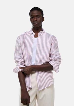 Jeune homme portant une chemise à boutons à rayures verticales blanches et roses par-dessus un t-shirt blanc et un pantalon crème, ajustant une manche retroussée.