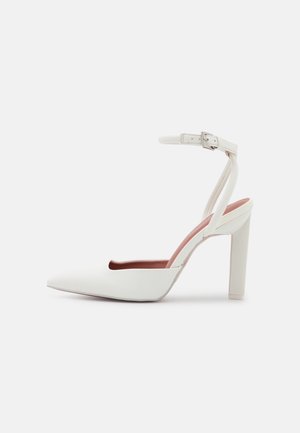 Chaussure à talon haut blanche avec bout pointu, bride de cheville avec boucle et talon bloc, présentée en profil latéral sur fond blanc.