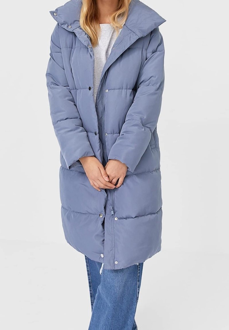 Femme portant un long manteau d'hiver rembourré bleu clair par-dessus un jean et une chemise blanche, debout avec les mains jointes devant elle.