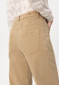 Beige cotton trousers featuring a smooth texture, five-pocket design, and a back patch with branding. Shaping is straight-cut with a subtle sheen.