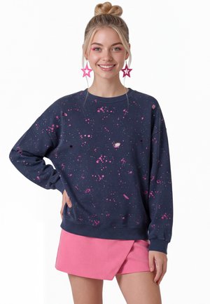 Ung kvinde med blondt hår sat op i en knold iført pink stjerneøreringe, en marineblå sweatshirt med pink pletter og et pink skørt, smilende med hånden på hoften.