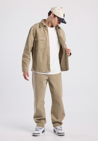 Giacca di cotone beige con tasche frontali, indossata sopra una maglietta bianca. Pantaloni di cotone beige e sneakers con dettagli grigi completano l'outfit.