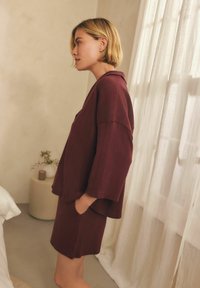 Completo da loungewear bordeaux composto da un giubbotto a vestibilitàampia con maniche a tre quarti e pantaloni corti abbinati, entrambi realizzati in tessuto testurizzato.