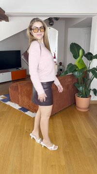 Roosa kudum-sviiter, pruun nahkne mini-seelik ja valged kontsakingad. Ruumi kaunistavad roheline taim ja oranž mööbel. Puitpõrand.