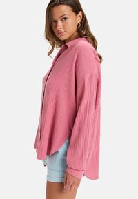 Helles rosa, strukturiertes Hemd mit langen Ärmeln; hat einen lockeren Schnitt und einen abgerundeten Saum. Kombiniert mit hellen Denim-Shorts.