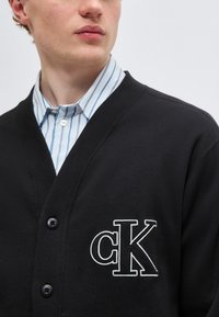 Νέος άνδρας που φοράει μαύρη ζακέτα με κουμπιά και λευκό λογότυπο "cK" πάνω από μπλε και λευκό ριγέ πουκάμισο, κοιτάζοντας προς τα πλάγια.