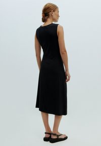 Schwarzes, ärmelloses Kleid mit leicht ausgestelltem Saum, aus glattem Stoff gefertigt, mit einem Detail am Rückenausschnitt. Getragen mit flachen, schwarzen Sandalen.