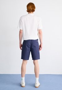 T-shirt bianca in cotone, maniche corte, vestibilità comoda. Pantaloni corti blu navy con due tasche posteriori e un piccolo logo. Calzature bianche con dettagli verdi.