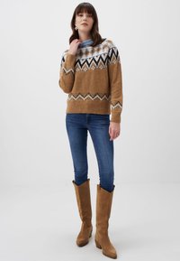 Maglione a maglia in un ricco color sabbia con motivi geometrici neri, bianchi e blu. Abbinato a jeans blu e stivali al ginocchio color sabbia.