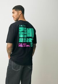 Tricou negru din bumbac, cu text îndrăzneț în verde și roz pe spate, cu un design inspirat de "Squid." Croială clasică, cu mâneci scurte.