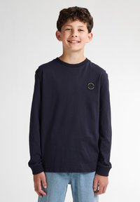 Camiseta de manga larga en algodón de color navy con cuello redondo y puños acanalados, que presenta un pequeño parche con el logo en el pecho. Lleva puesta con jeans de color azul claro.