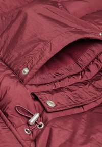 Chaqueta acolchada burdeos con una cubierta texturizada, que presenta botones de presión plateados, cordón elástico y un forro interior suave de contraste.