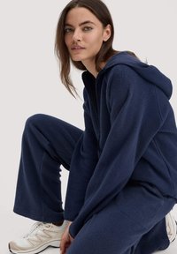 Marineblaue Fleece-Hoodie und passende Hose. Weicher Stoff, lockere Passform, ausgestattet mit einer Kapuze. Weiße Sportschuhe mit beigen Akzenten.
