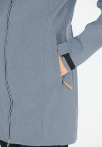 Gray fabric jacket with a zip closure, side pockets, and a contrasting black Velcro cuff. Features a subtle textured pattern and minimalist design.