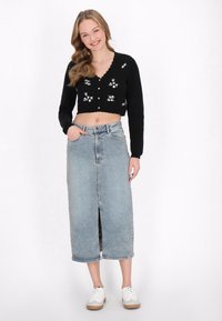 Cárdigan negro corto con bordados florales, combinado con una falda maxi de mezclilla azul clara con una abertura frontal. Zapatillas blancas completan el conjunto.