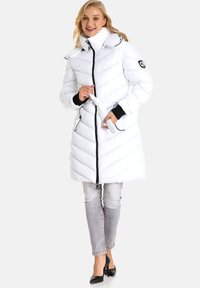 Weißer, puffiger Wintermantel mit Zickzacksteppung, hohem Kragen, schwarzem Reißverschluss und Gürteldetail. Kombiniert mit hellgrauen Jeans und schwarzen High Heels.