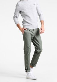 Světle šedý svetr s žebrovanými lemy, kombinovaný s ohrnutými olivově zelenými kalhotami. Bílé tenisky doplňují outfit a zdůrazňují uvolněný styl.