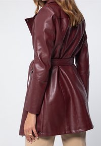 Donna che indossa una giacca di pelle rossa scura con cintura e maniche lunghe, mostrata di spalle e di lato, abbinata a pantaloni beige.