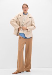 Giacca in misto lana beige con colletto ampio, abbinata a un maglione lavorato a maglia crema e una camicia blu, completata da pantaloni a gamba larga marroni e tacchi.