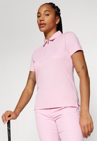 Camisa polo rosa com gola com fecho, mangas curtas e tecido texturizado. Usada com calças rosas combinando e segurando um taco de golfe preto.