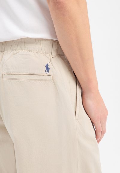 Pantalon beige avec taille élastique et logo joueur de polo brodé en bleu, porté avec une chemise blanche, montrant les détails des poches latérales et arrière.